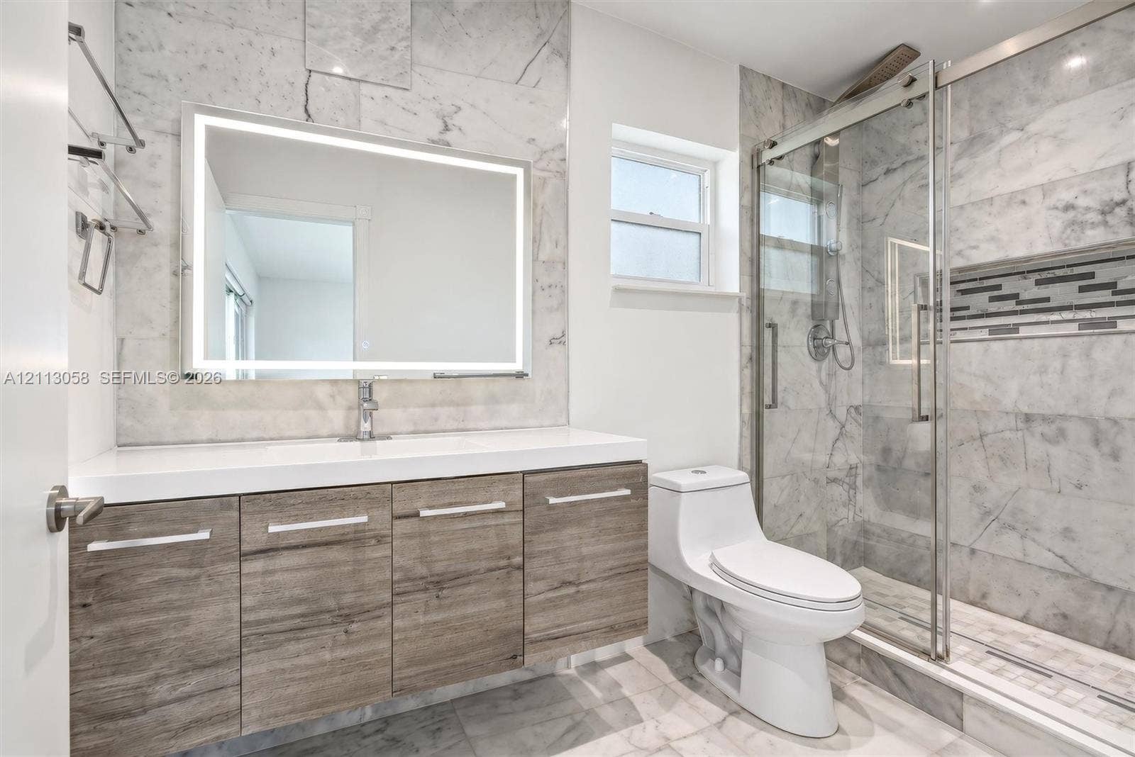 1980 NW 33rd Court, Oakland Park, FL 33309 Modern bathroom featuring wood-finish vanity cabinetry, a white countertop with an integrated sink, floor-to-ceiling stone-tiled walls, a backlit mirror, and a glass-enclosed shower with a decorative mosaic tile accent