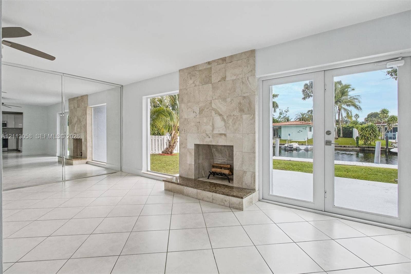 1980 NW 33rd Court, Oakland Park, FL 33309 Neutral tile flooring, floor-to-ceiling mirrored wall panels, stone-tiled fireplace with a raised hearth, and double glass doors opening to a waterfront exterior