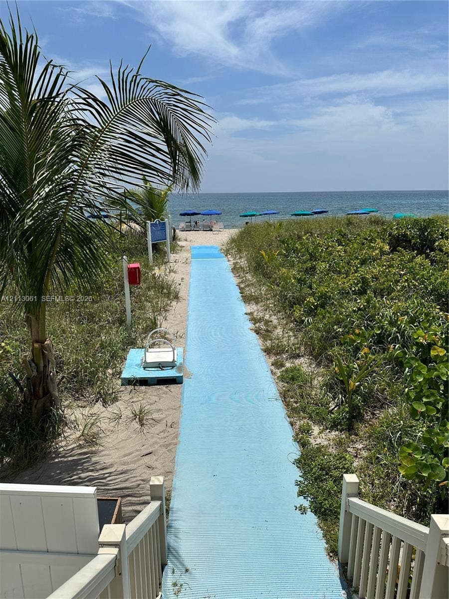 Direct beach access featuring a paved walkway, sandy shoreline, mature coastal vegetation, and a wooden staircase