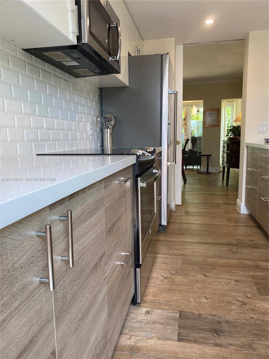 Kitchen featuring wood-finish cabinetry with bar pulls, white solid-surface countertops, a white subway tile backsplash, stainless steel appliances, and wood-finish flooring
