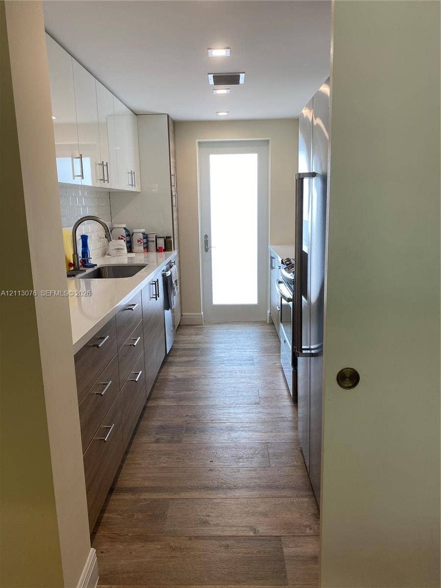 Galley kitchen featuring wood-finish cabinetry, white slab countertops, stainless steel appliances, wood-finish flooring, and a glass-paneled exterior door