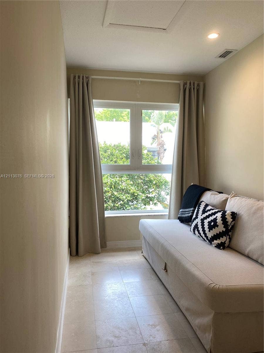 Neutral-toned interior space featuring light-colored stone flooring, recessed ceiling lighting, and a large multi-pane window with natural exterior greenery