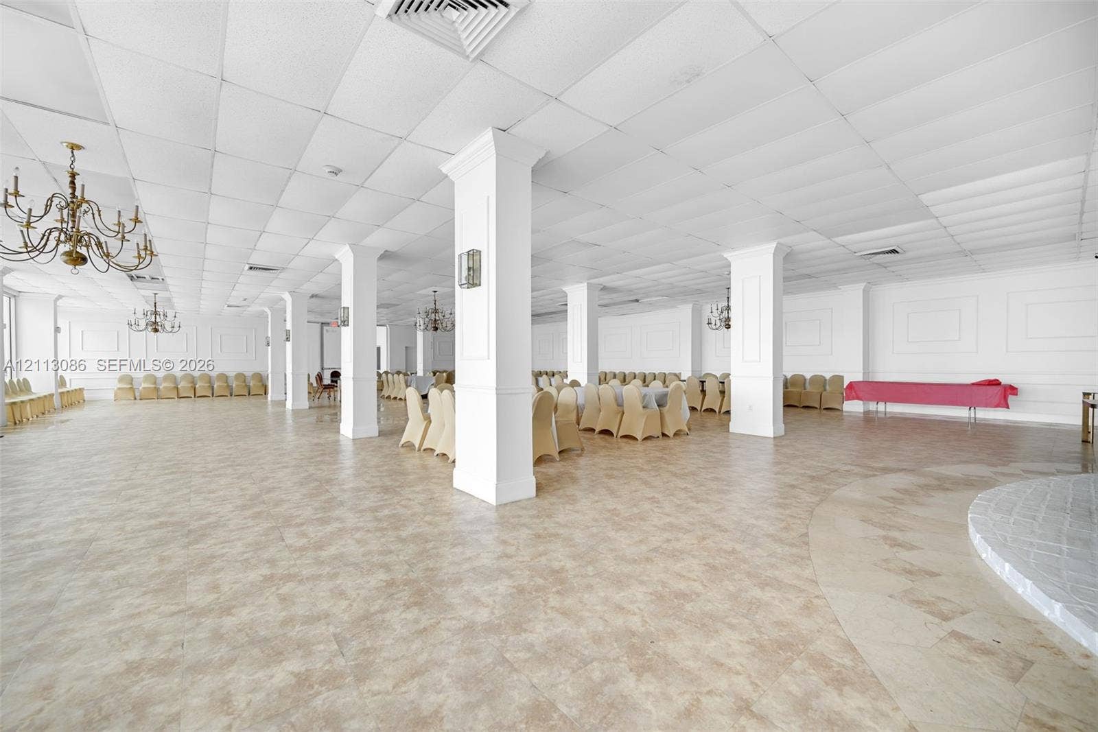 7135 Collins Avenue, #1632, Miami Beach, FL 33141 Expansive open-concept hall featuring a dropped acoustic tile ceiling, structural columns with wainscoting detail, wall-mounted light fixtures, tile flooring, and multiple ornate chandeliers