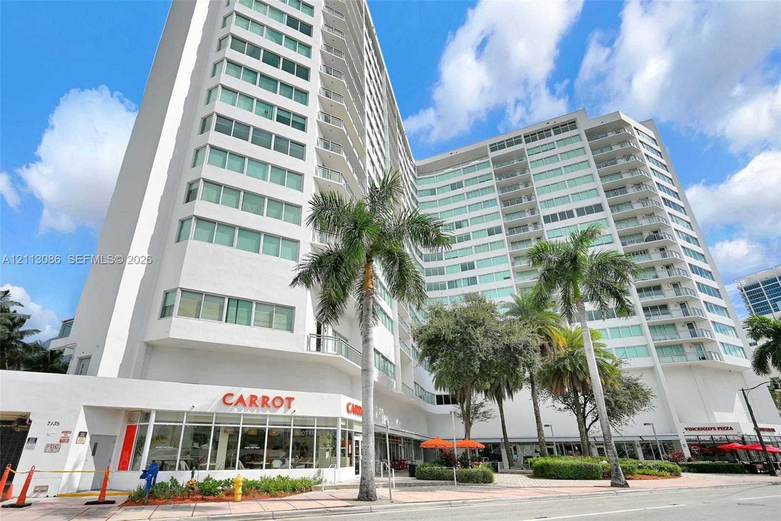7135 Collins Avenue, #1632, Miami Beach, FL 33141 High-rise residential building featuring a white facade, extensive glass window panels, wrap-around private balconies, and ground-level commercial storefronts