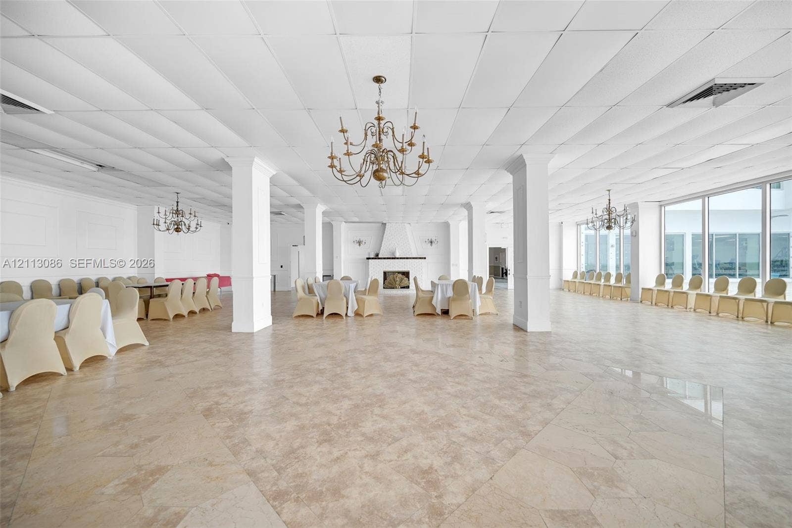 7135 Collins Avenue, #1632, Miami Beach, FL 33141 Expansive open-concept hall featuring large-format tile flooring, a central fireplace with a decorative mantel, structural columns, ornate multi-arm chandeliers, and floor-to-ceiling windows