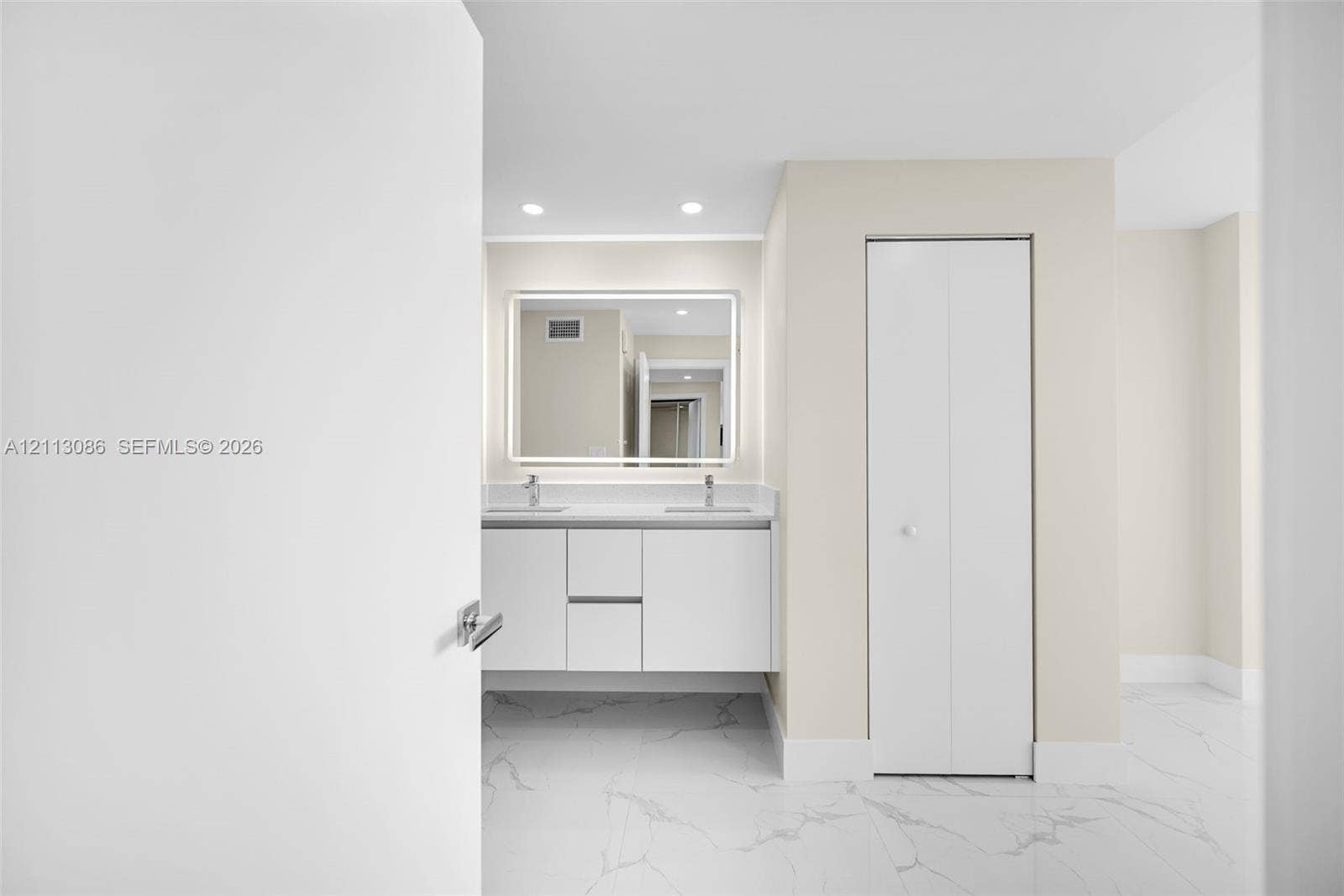 7135 Collins Avenue, #1632, Miami Beach, FL 33141 Floating vanity with double under-mount sinks, integrated stone-surface countertop, LED-backlit mirror, and white bi-fold closet doors, finished with large-format light-toned stone-patterned flooring