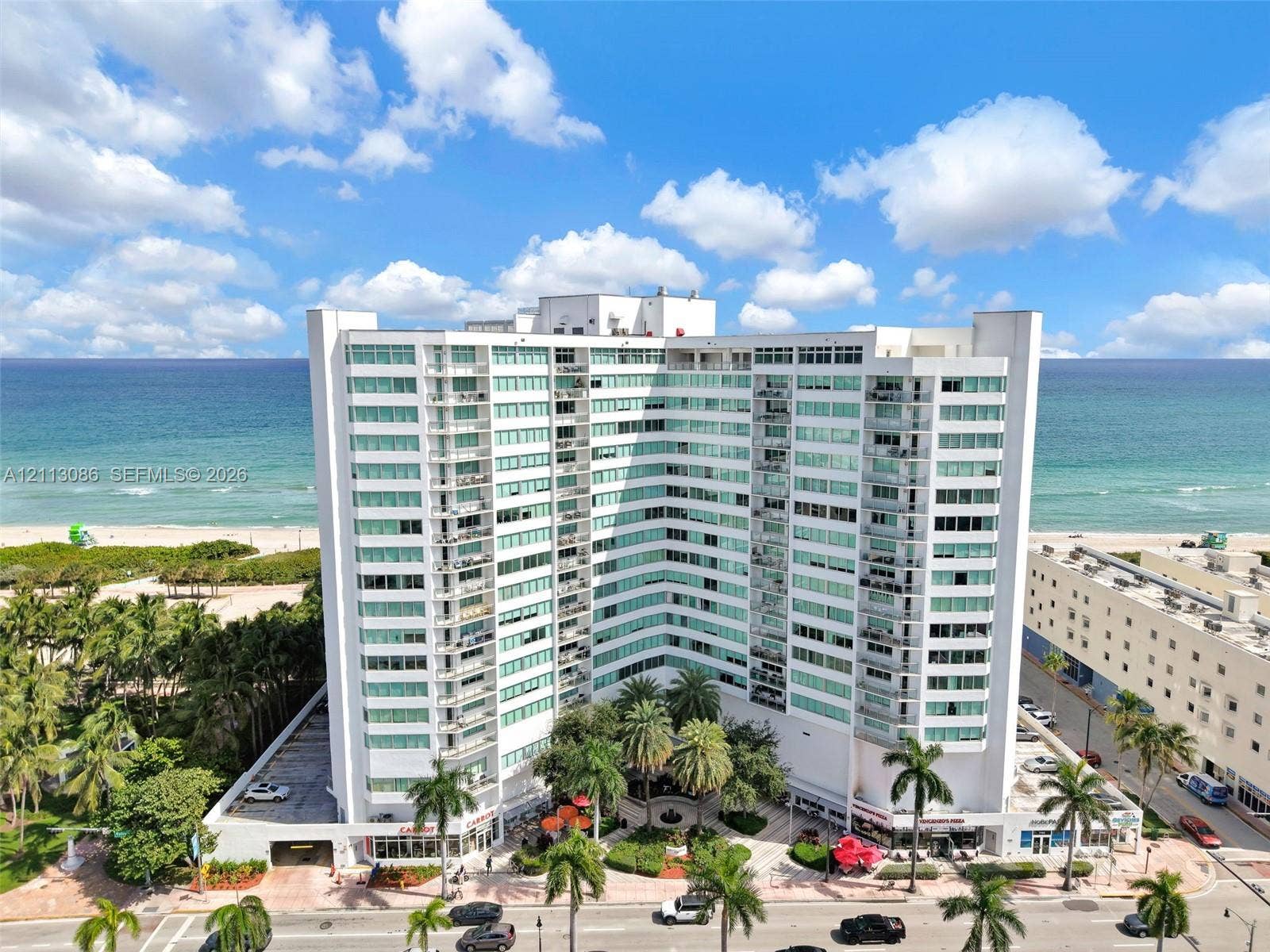 7135 Collins Avenue, #1632, Miami Beach, FL 33141 High-rise building featuring a white facade, multiple stacked balconies, floor-to-ceiling glass windows, ground-level covered parking, and an established landscaped courtyard
