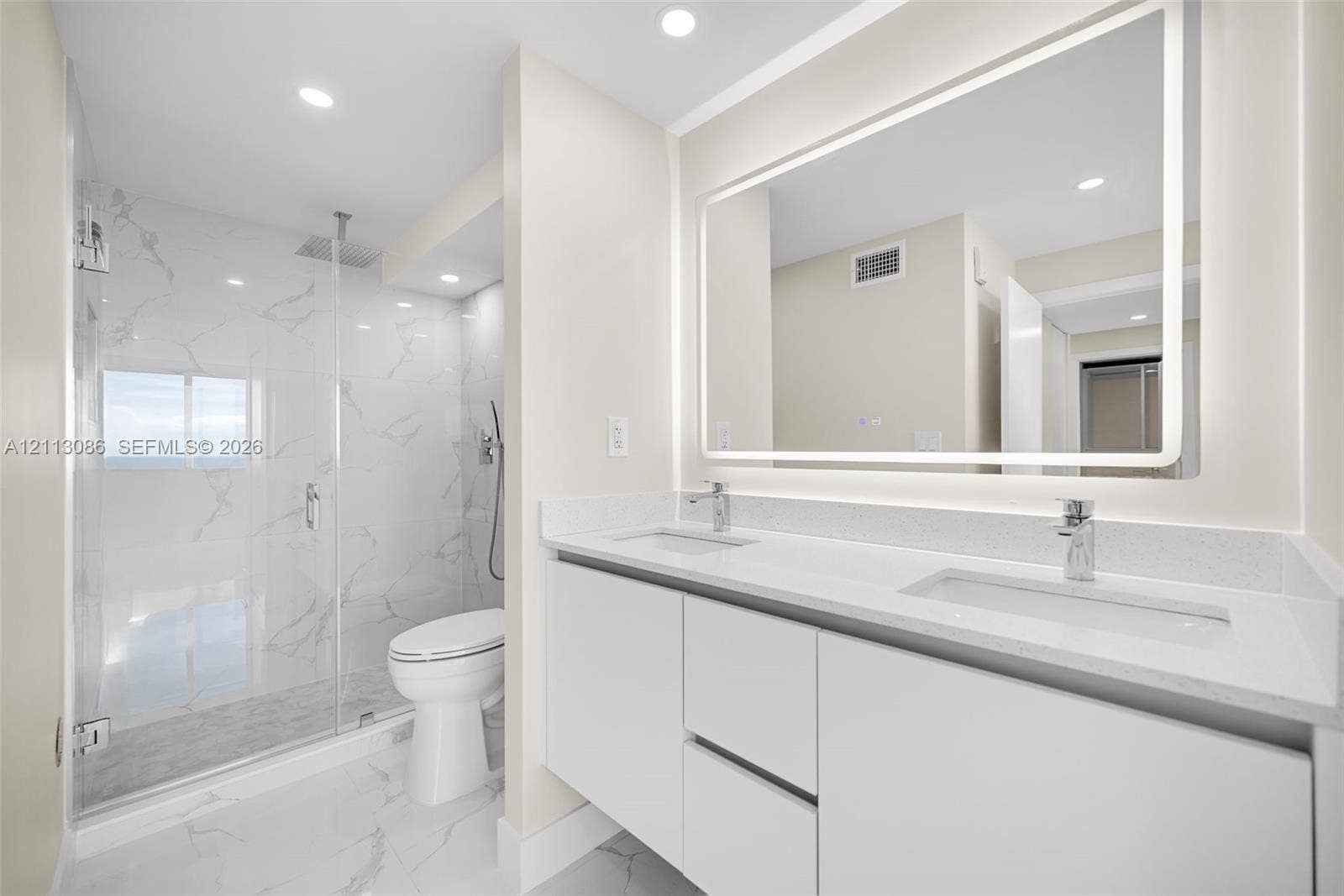 7135 Collins Avenue, #1632, Miami Beach, FL 33141 Dual-sink vanity with white cabinetry and solid surface countertop, backlit vanity mirror, walk-in glass shower enclosure featuring rainfall shower head and floor-to-ceiling stone-finish tile surround, and coordinating stone-finish flooring