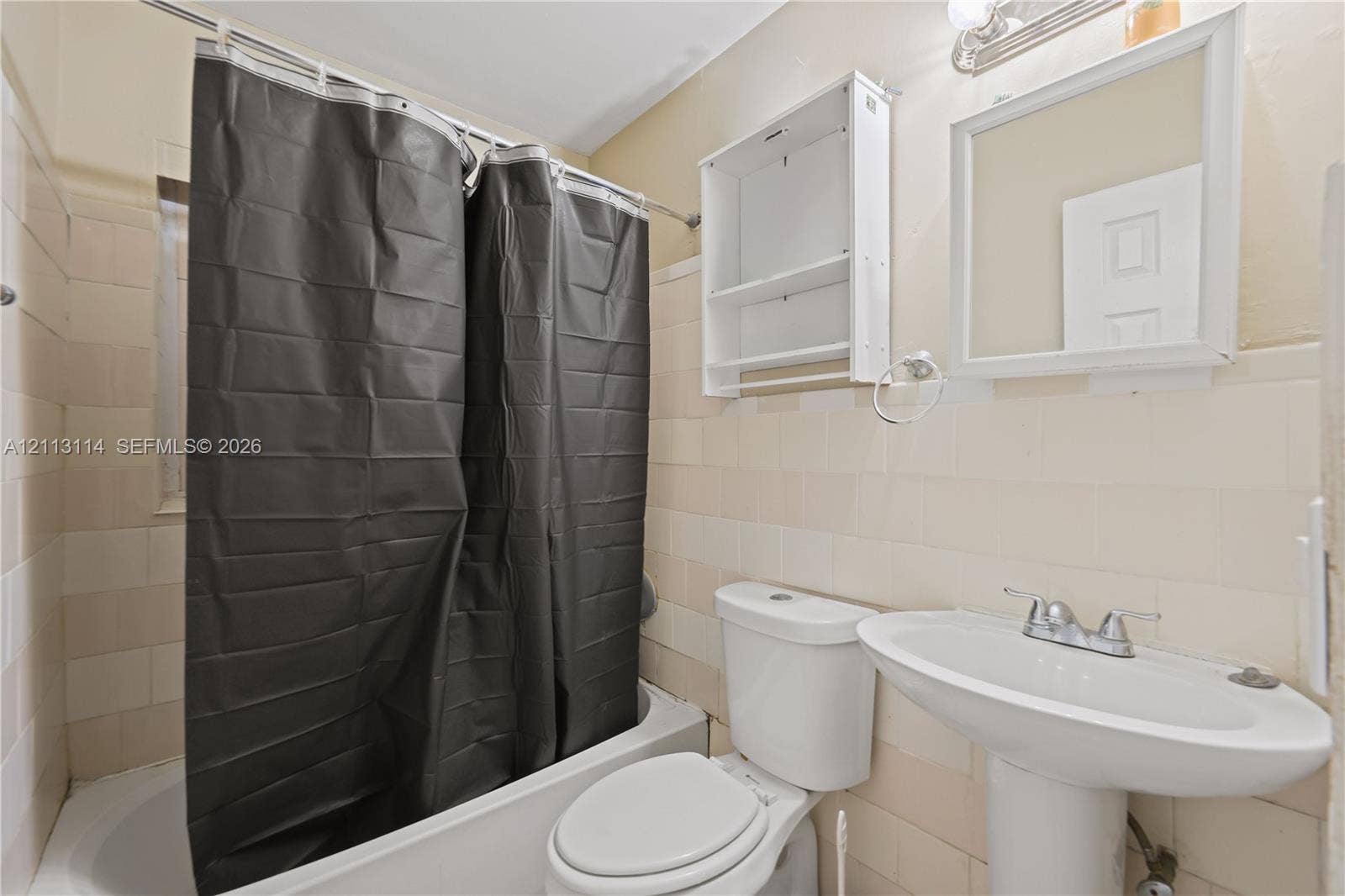 Full bathroom featuring a white pedestal sink, a bathtub with wall-mounted plumbing fixtures, neutral ceramic wall tile, a wall-mounted storage cabinet, and a framed vanity mirror