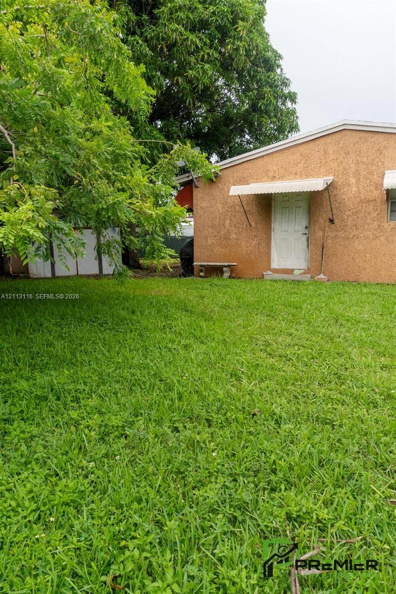 4341 NW 178th Drive, Miami Gardens, FL 33055 Rear yard featuring a grass lawn, mature trees, textured stucco exterior, and a metal door awning