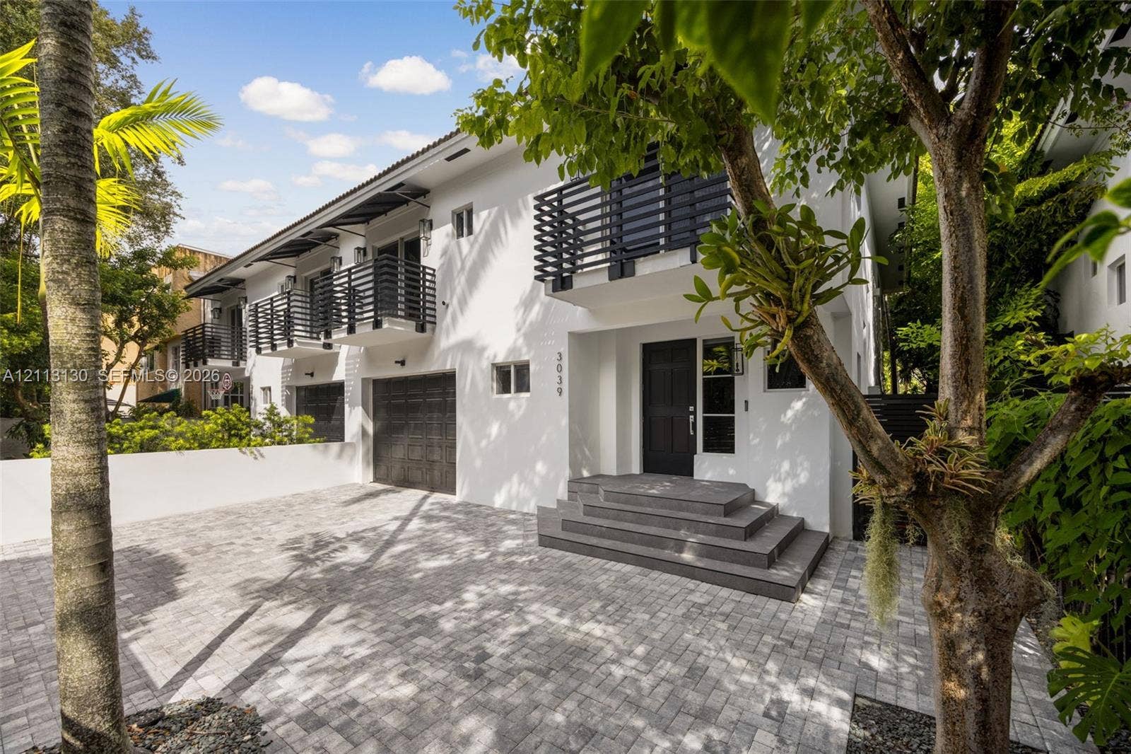 Stucco exterior featuring dual-level black metal balconies, a paved stone driveway, an attached garage, and tiered concrete front entry steps