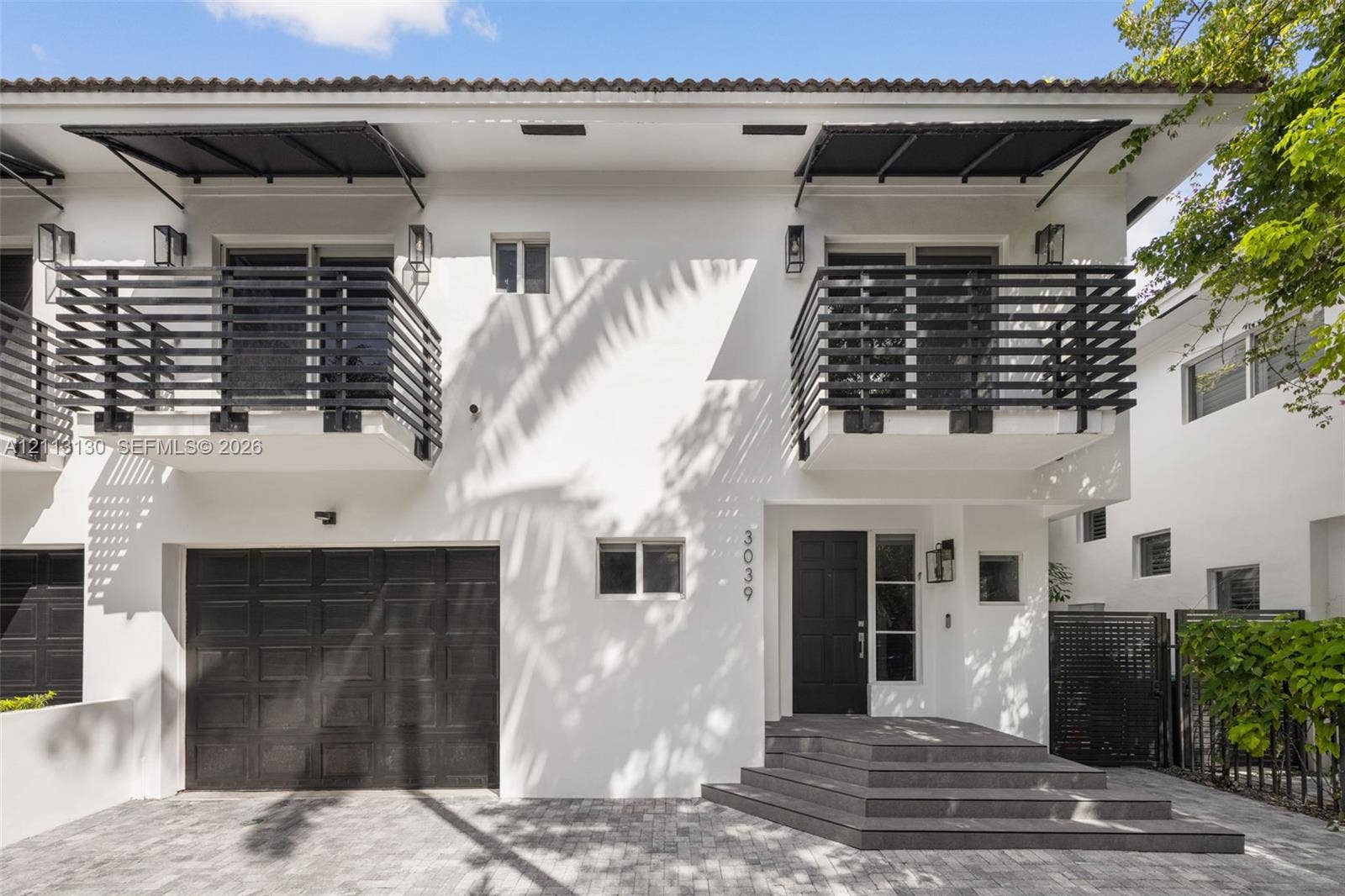 Two-story stucco exterior featuring horizontal black metal balcony railings, a multi-paneled garage door, geometric entry steps, and matching black metal awnings