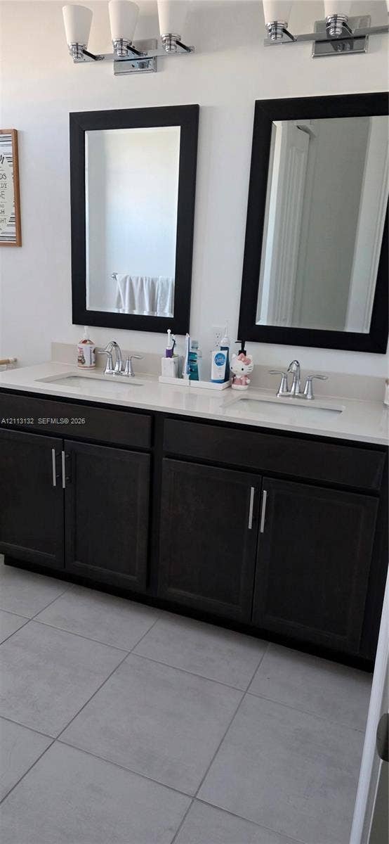 16474 Sea Turtle Place, Westlake, Westlake, FL 33470 Double-sink vanity featuring dark wood cabinetry, solid-surface countertop with integrated basins, chrome hardware, matching framed mirrors, and dual-light wall fixtures over large-format tile flooring