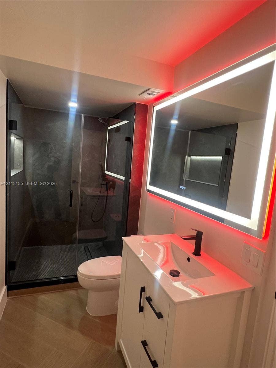 1600 S Ocean Drive, #3A, Hollywood, FL 33019 Bathroom featuring a vanity with white cabinetry and matte black hardware, integrated sink with a matte black faucet, wall-mounted LED-backlit mirror, glass-enclosed shower with dark tile surround, and tile flooring