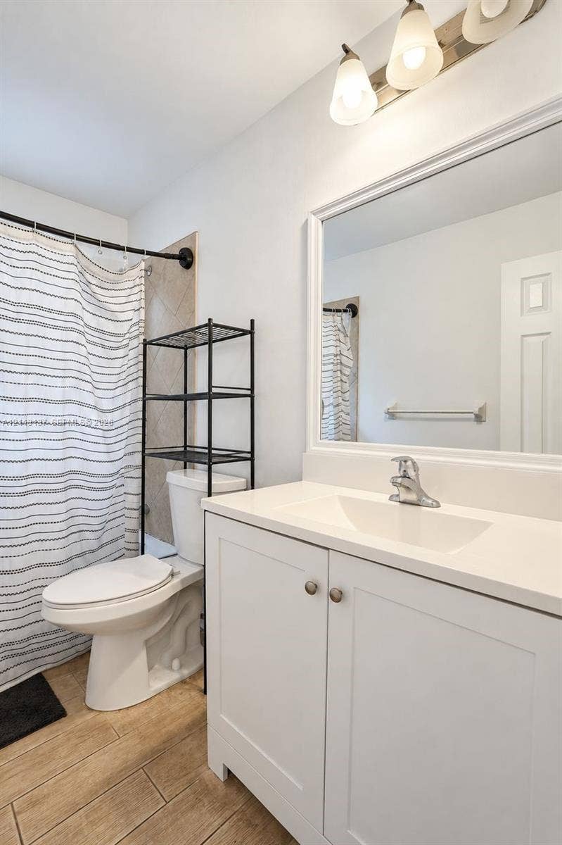 10884 Royal Palm Boulevard, #10884, Coral Springs, FL 33065 White vanity with a solid-surface countertop and integrated sink, framed wall mirror, three-light vanity fixture, and wood-finish flooring