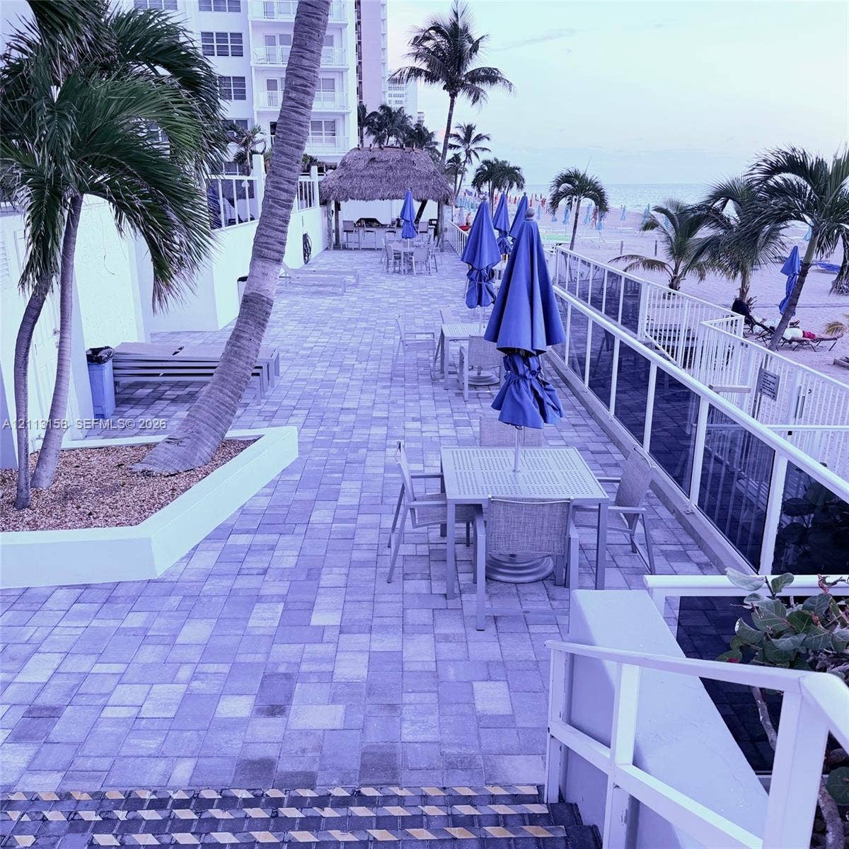 Paver patio terrace featuring a built-in thatched-roof tiki hut, white metal perimeter railing, and integrated palm tree planters