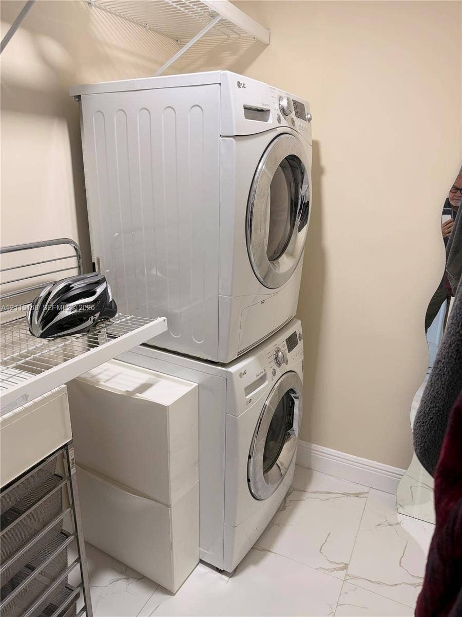 Stacked front-load laundry appliances, white stone-finish tile flooring, and integrated white wire shelving