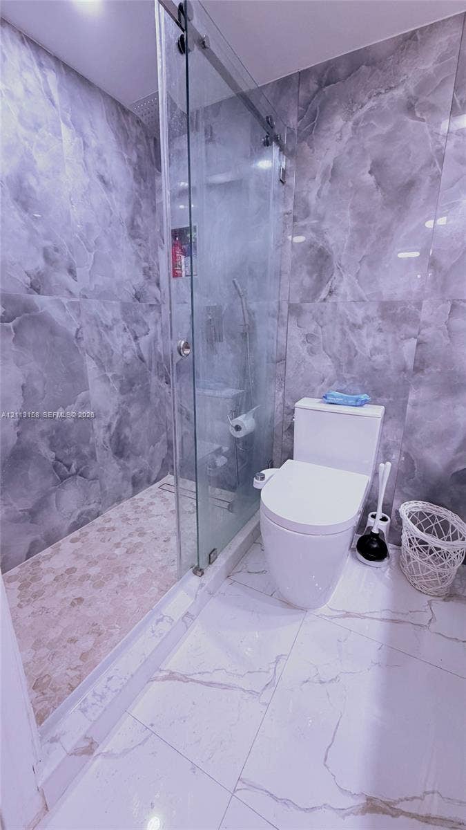 Walk-in shower with a sliding glass door, floor-to-ceiling gray-toned wall tiling, pebble-finish shower flooring, and oversized white floor tile with gray veining