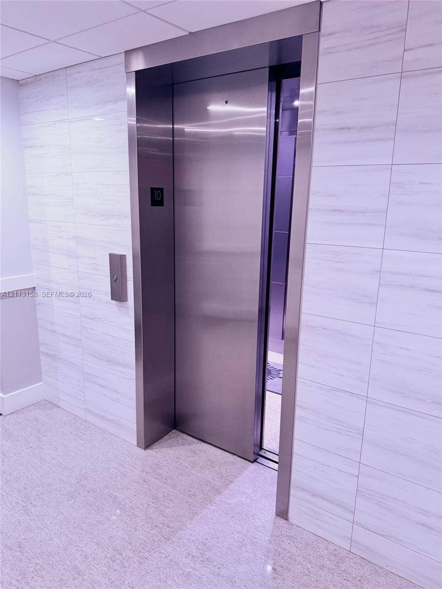 Stainless steel elevator with a brushed metal frame, surrounded by large-format, horizontal-vein tile wall cladding and a polished stone-finish floor