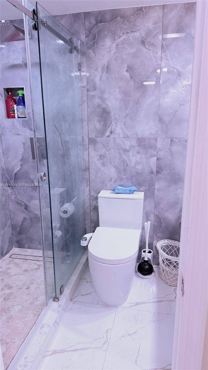 Walk-in shower with a sliding glass door, linear drain, built-in wall niche, and large-format gray stone-finish wall tile, complemented by white large-format stone-finish floor tile and a dual-flush toilet