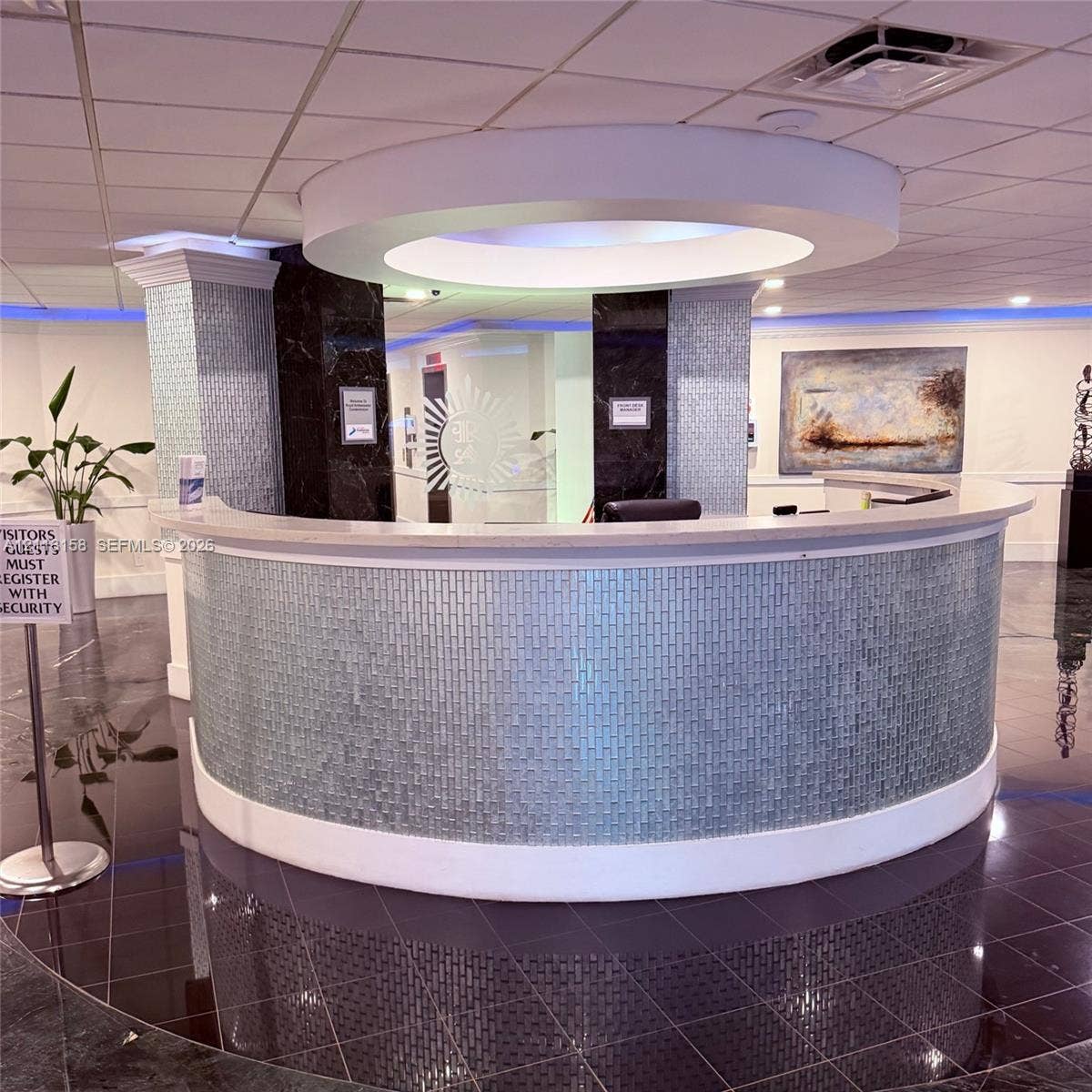 Curved reception desk featuring a light-toned solid surface countertop and glass tile facade, suspended circular ceiling soffit with integrated lighting, and high-gloss dark tile flooring