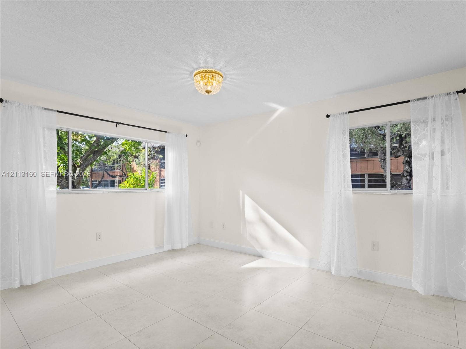 Neutral-toned interior featuring large-format tile flooring, dual windows with black curtain rods, and a central flush-mount ceiling fixture