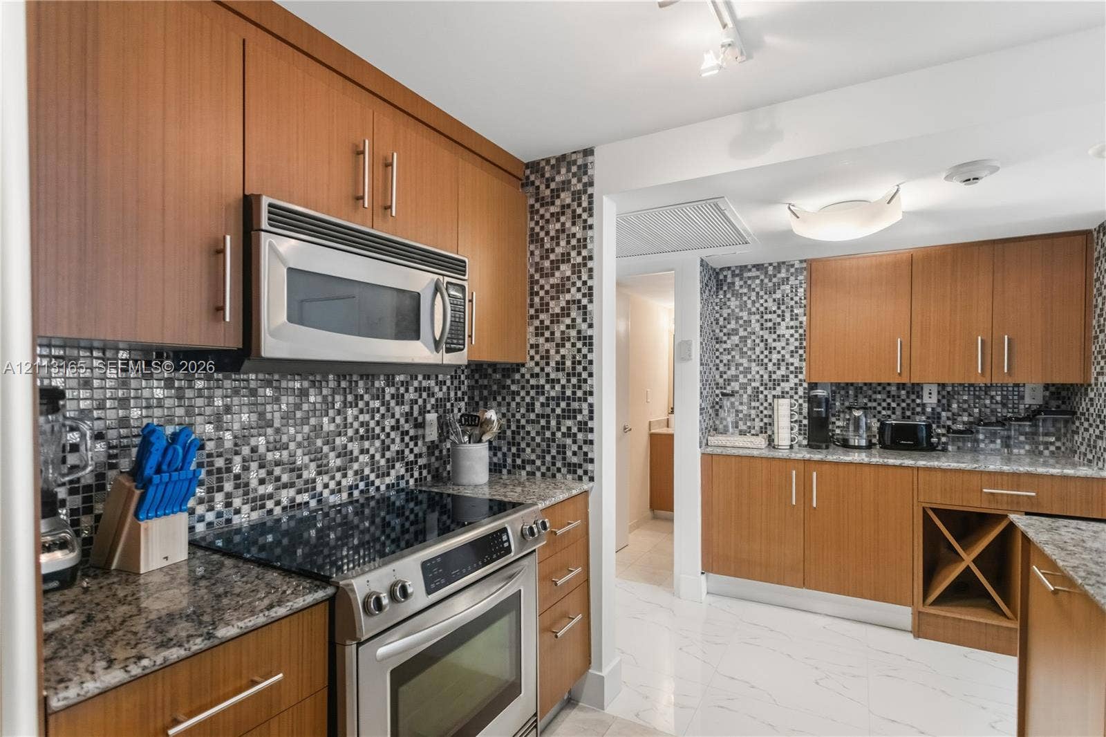 10275 Collins Avenue, #229, Bal Harbour, FL 33154 Kitchen featuring wood-finish cabinetry, stone countertops, mosaic tile backsplash, built-in wine rack, and stainless steel appliances