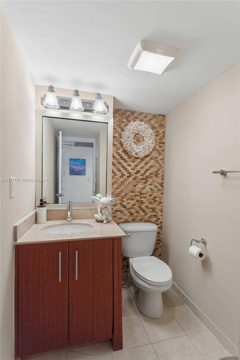 10275 Collins Avenue, #229, Bal Harbour, FL 33154 Vanity with a solid-surface countertop and integrated sink, wood-finish cabinetry, mosaic-tiled accent wall, three-light fixture, and ceramic tile flooring