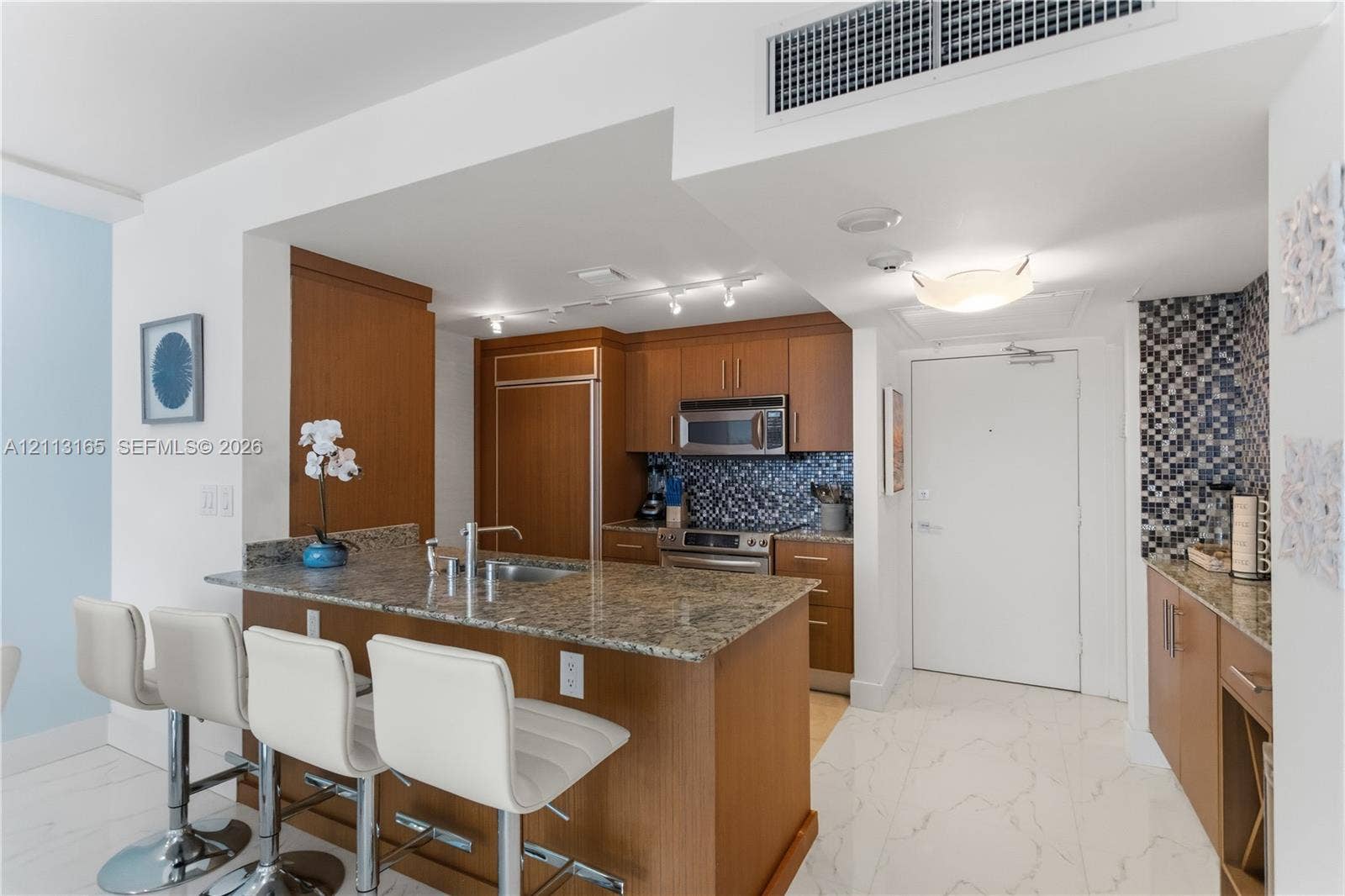 10275 Collins Avenue, #229, Bal Harbour, FL 33154 Kitchen featuring wood-finish cabinetry, stone countertops, a mosaic tile backsplash, stainless steel appliances, and large-format tile flooring