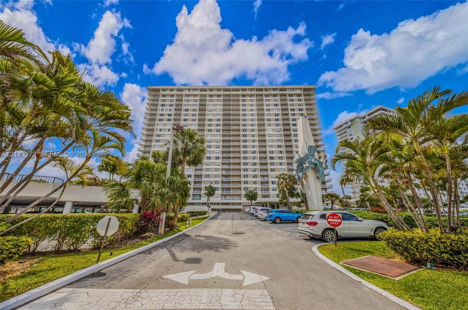 300 Bayview Drive, #1614, Sunny Isles Beach, FL 33160 High-rise residential exterior featuring a multi-story facade, paved surface parking, integrated landscaping, and sculptural monument