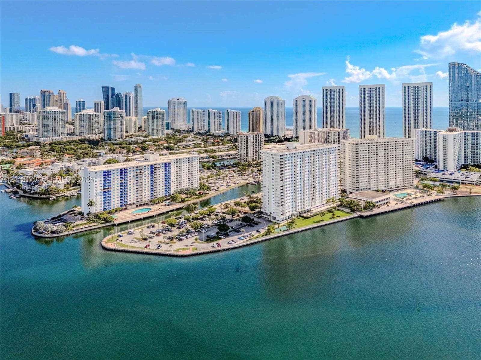 300 Bayview Drive, #1614, Sunny Isles Beach, FL 33160 Waterfront aerial perspective featuring multiple high-rise structures, paved parking areas, and an expansive bay shoreline