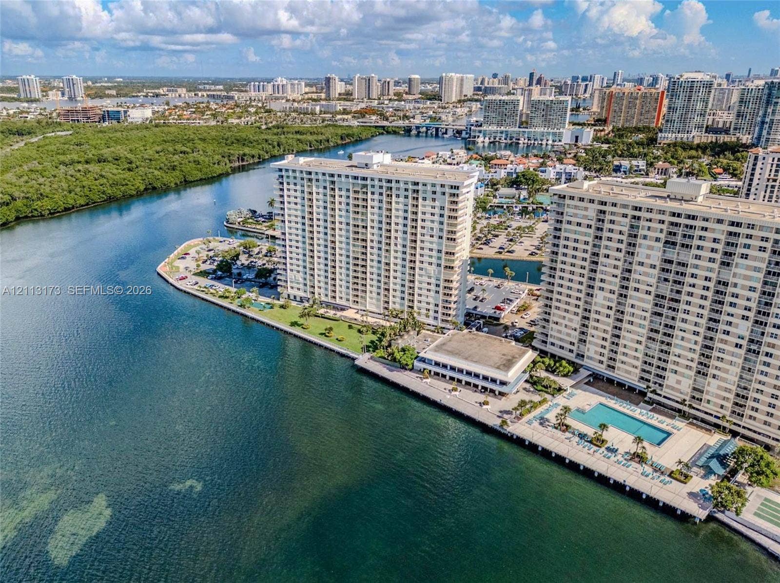 300 Bayview Drive, #1614, Sunny Isles Beach, FL 33160 High-rise waterfront complex featuring a rectangular swimming pool, concrete seawall, paved parking areas, and manicured green spaces