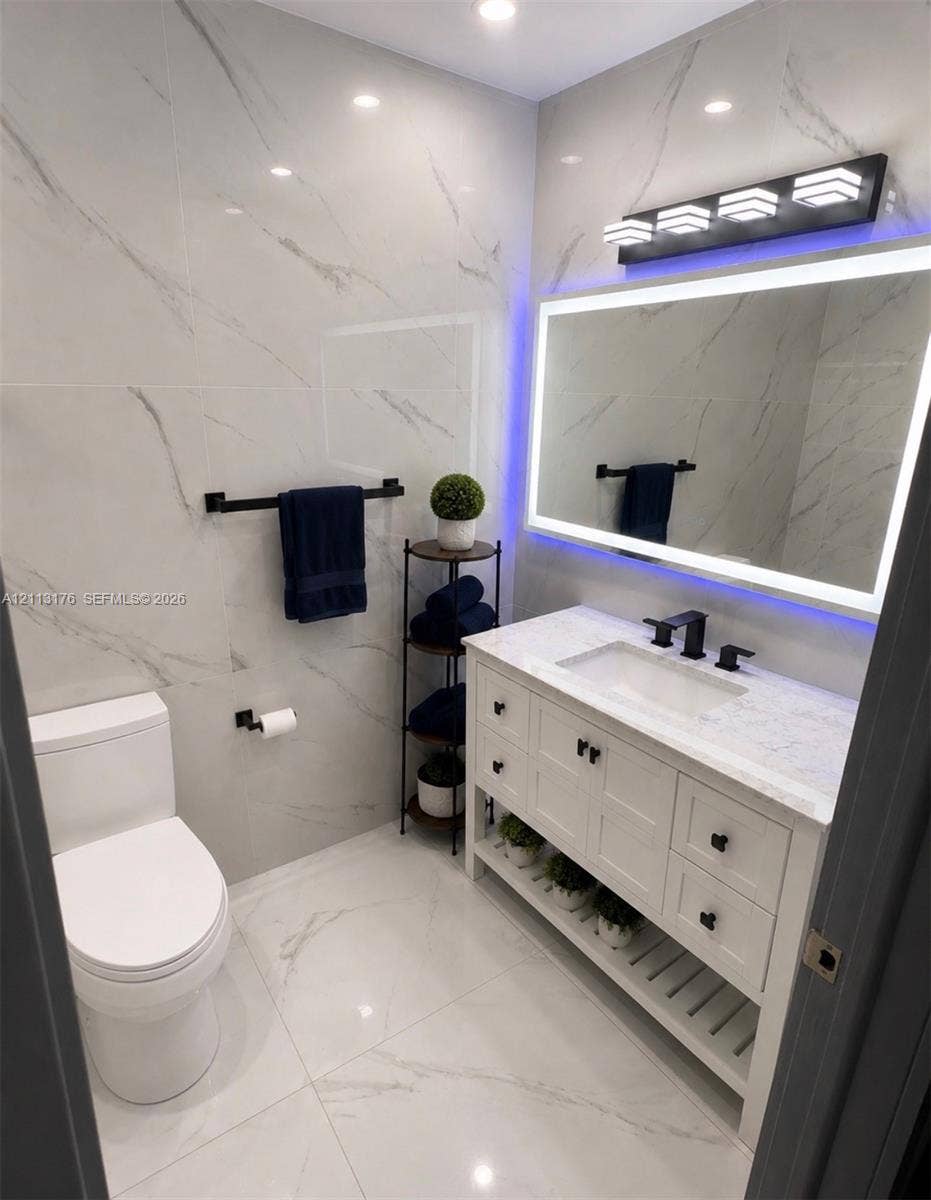 4391 NW 112th Avenue, Coral Springs, FL 33065 Bathroom featuring floor-to-ceiling large-format stone-patterned tile, white vanity with stone countertop, undermount sink with matte black widespread faucet, LED-backlit mirror, and matte black hardware