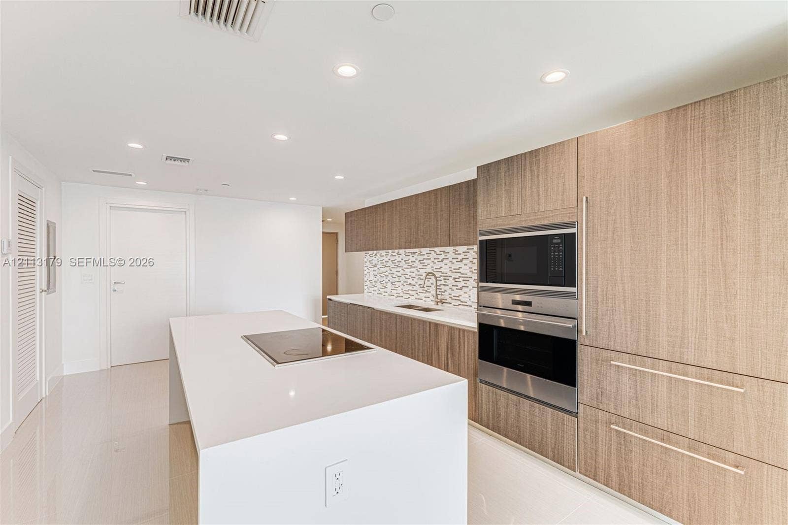 1150 102nd Street, #703, Bay Harbor Islands, FL 33154 Kitchen featuring wood-finish cabinetry, a white countertop island with an integrated cooktop, stainless steel wall-mounted oven and microwave, stone mosaic backsplash, and recessed lighting