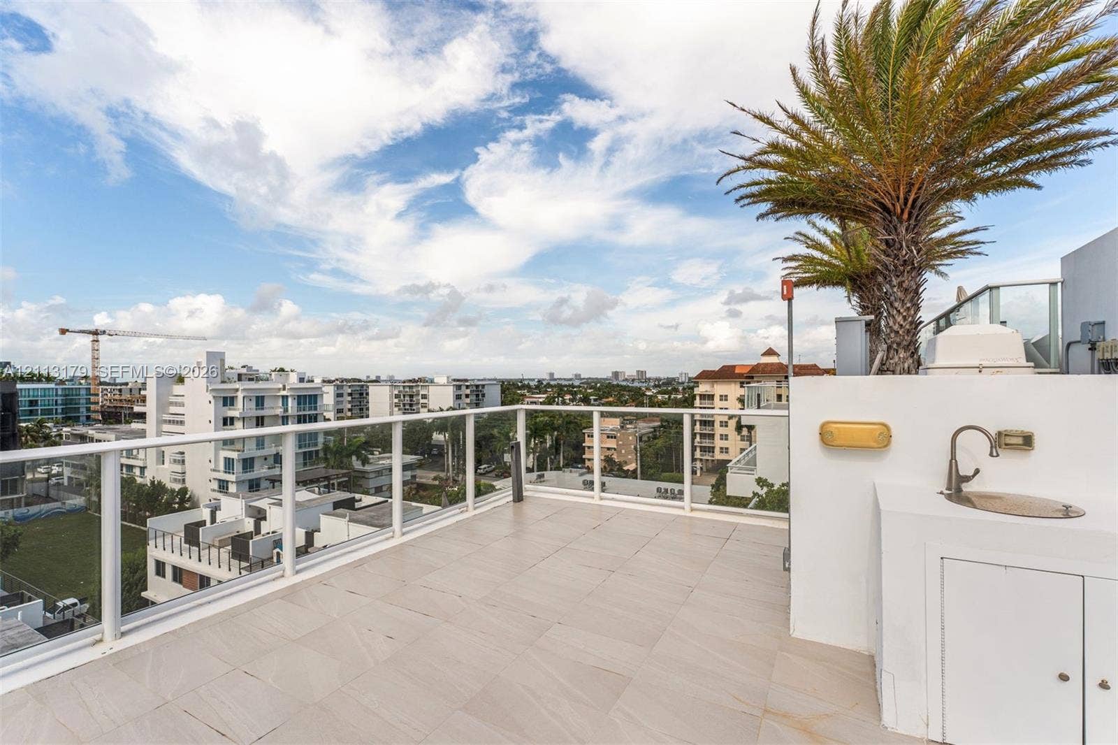 1150 102nd Street, #703, Bay Harbor Islands, FL 33154 Rooftop terrace featuring light-toned tile flooring, a white glass-paneled railing, integrated wet bar with a stainless steel sink, and mature palm trees