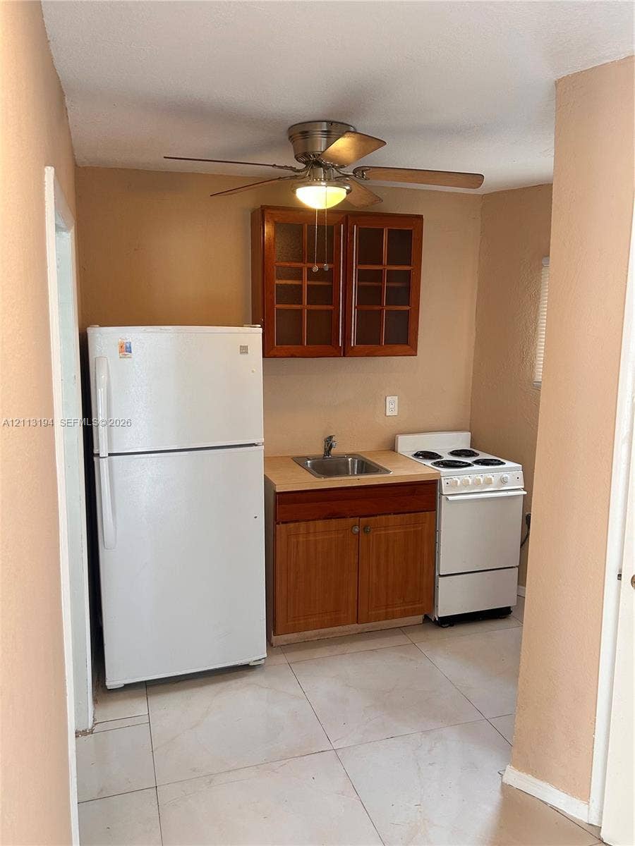 Kitchenette featuring light-toned tile flooring, wood cabinetry with glass-panel upper doors, a stainless steel sink, an electric range, and a ceiling-mounted light fixture with fan
