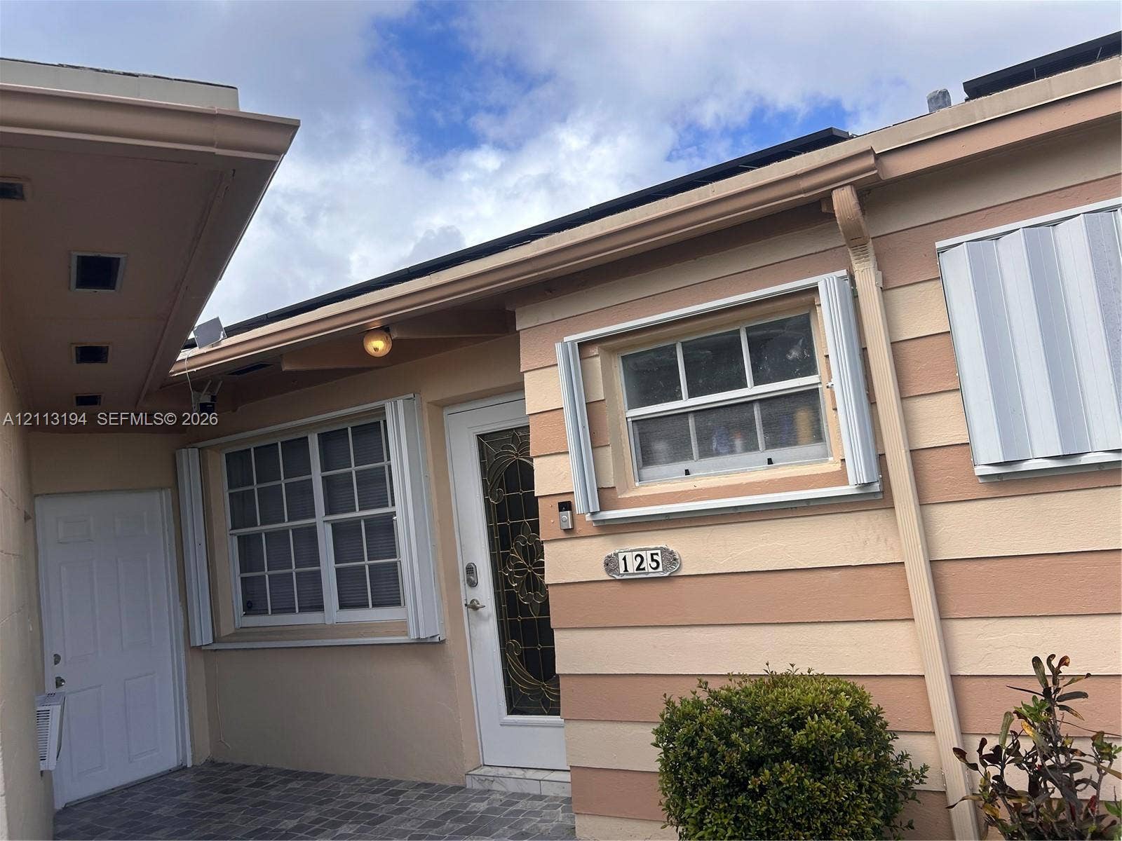 Exterior facade featuring horizontal two-tone siding, grid-patterned windows, decorative glass entry door, and stone-finish paved entry area