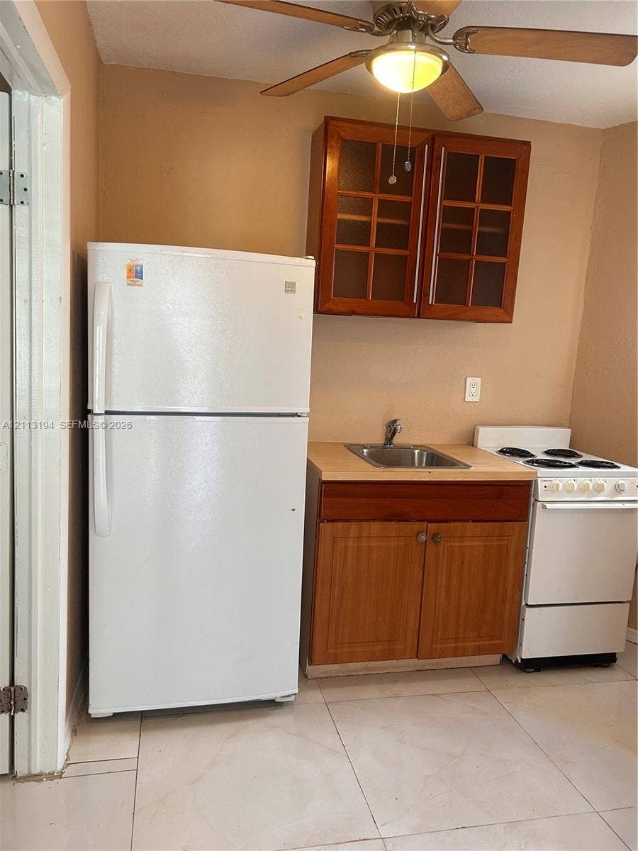Kitchenette featuring wood-finish cabinetry with glass-paneled upper doors, a stainless steel sink, a white refrigerator, a white electric range, and tile flooring