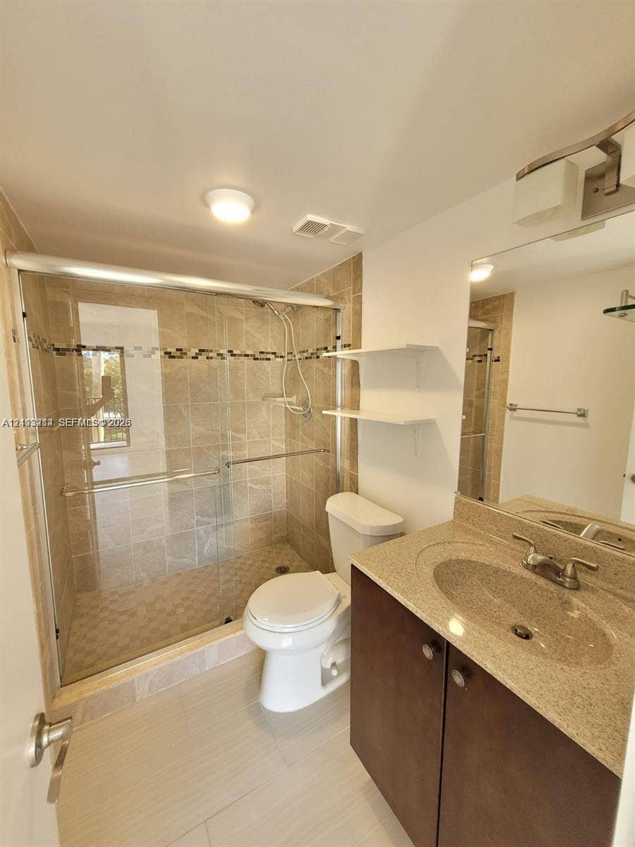 Bathroom featuring a vanity with a stone-finish countertop, under-mount sink, and dark cabinetry, alongside a walk-in shower with a sliding glass door, mosaic tile accents, and integrated wall shelving