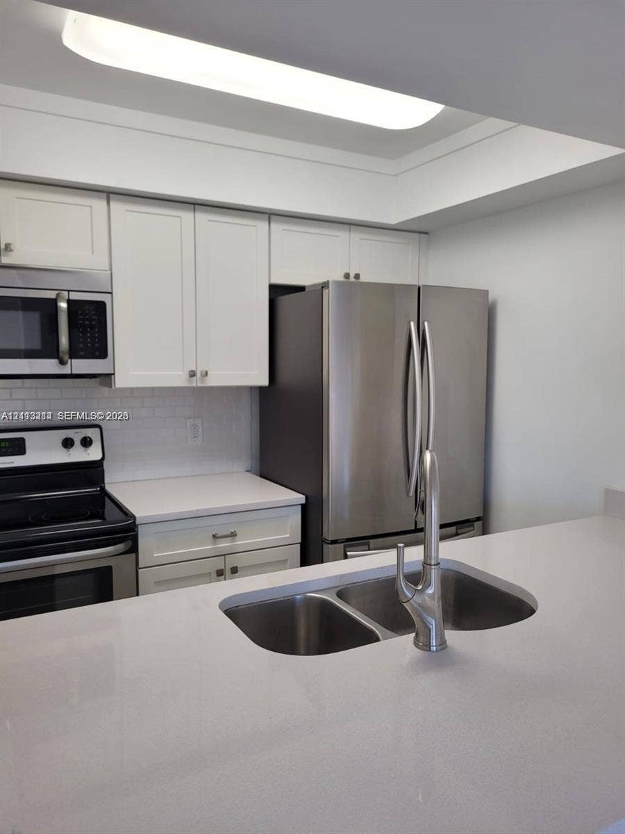 Kitchen featuring white shaker-style cabinetry, solid-surface countertops, subway tile backsplash, stainless steel appliances, and an undermount dual-basin sink with a gooseneck faucet