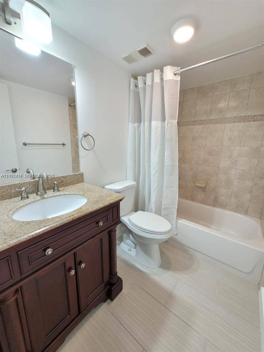 Bathroom featuring a dark wood-finish vanity, stone-surface countertop with an undermount sink, ceramic tile shower surround, and neutral-tone flooring