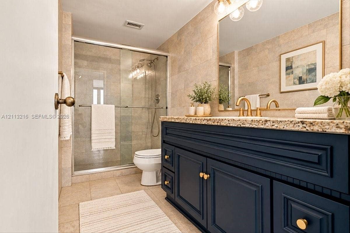 900 Bay Drive, #425, Miami Beach, FL 33141 Bathroom featuring navy blue cabinetry, speckled stone countertop, brass-tone hardware, large wall-mounted mirror, and a glass-enclosed shower with tiled surround