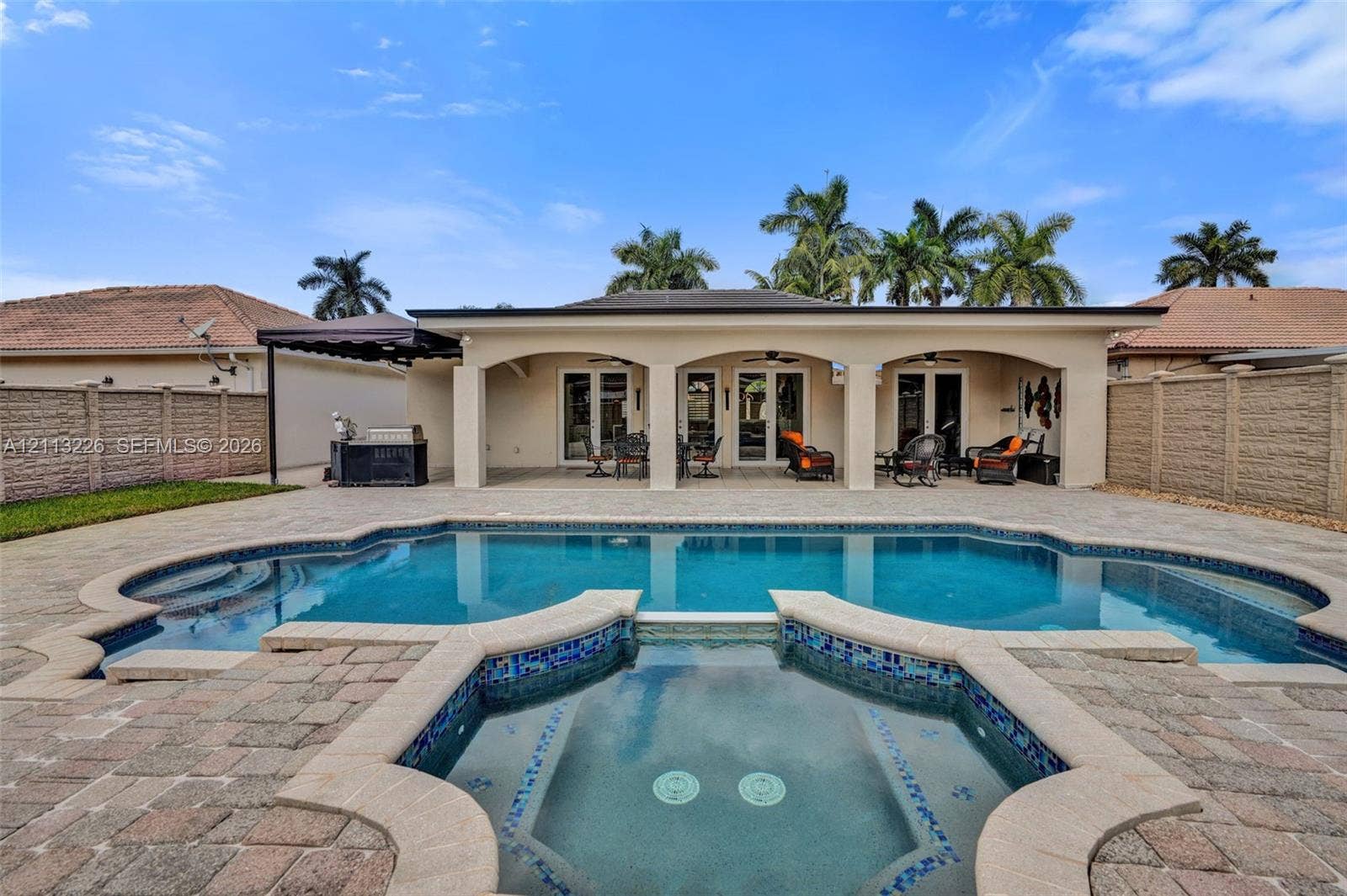 Backyard featuring an inground pool with an integrated spa, paved pool deck, covered patio with arched supports, and perimeter masonry fencing