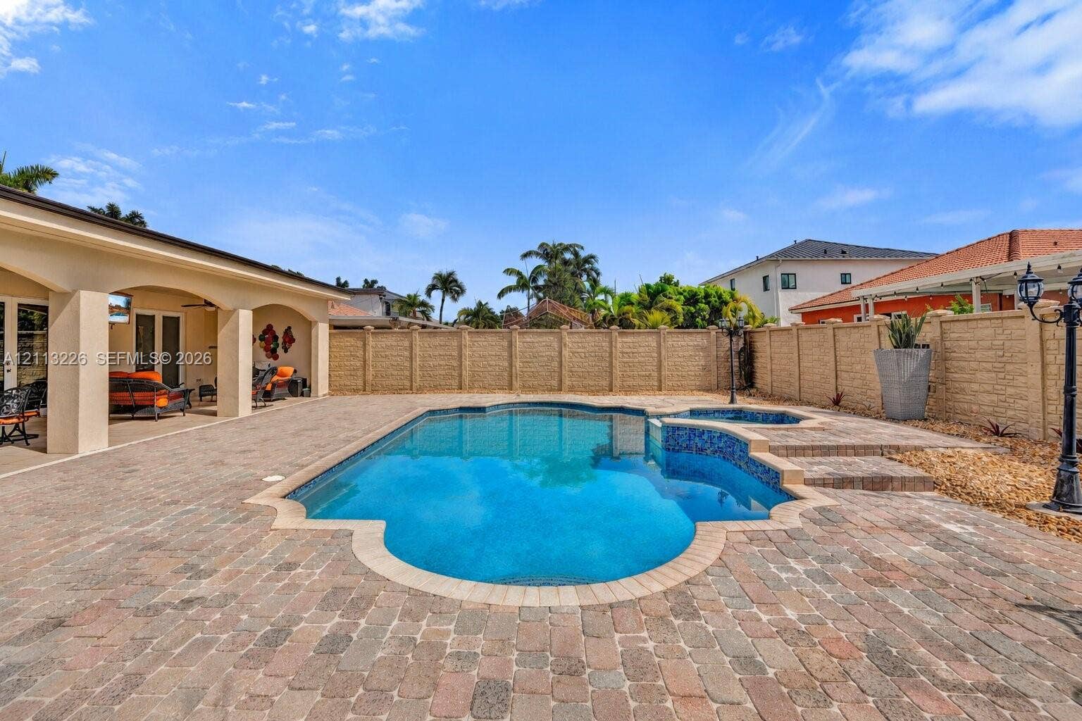 Rear patio featuring a covered loggia, paver-stone hardscaping, a custom-shaped swimming pool with decorative tile accents, an integrated raised spa, and perimeter masonry wall fencing