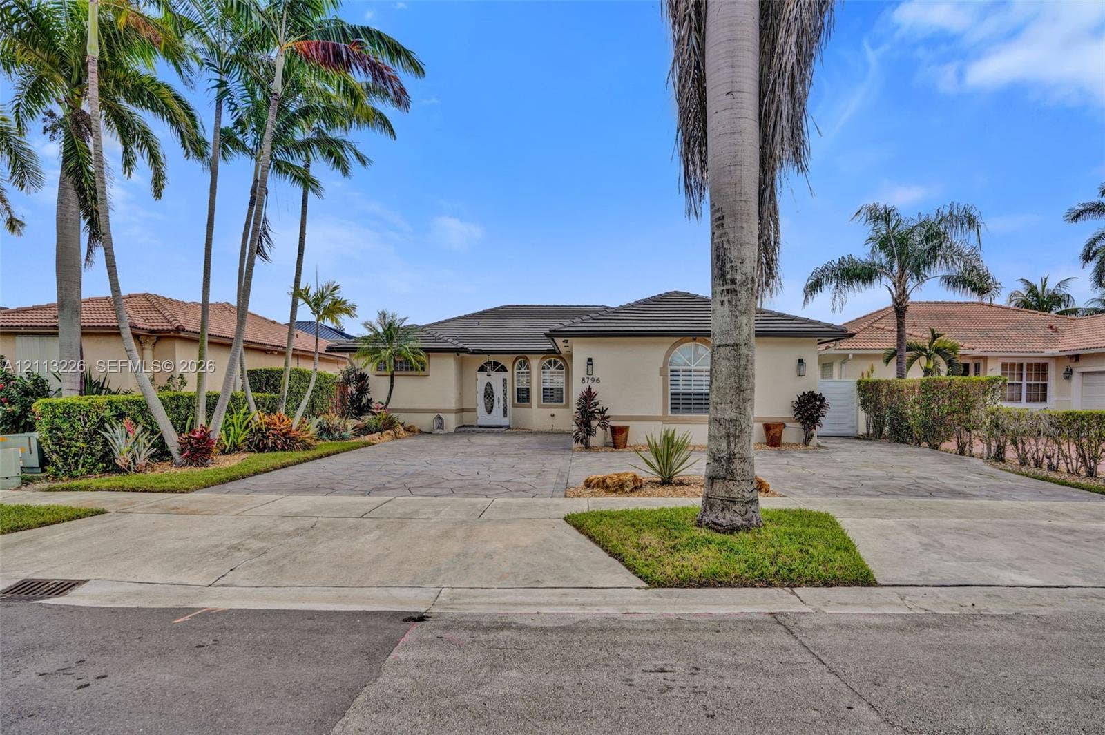 Single-story residence featuring a tile roof, light-toned stucco exterior, decorative front door with glass inlay, stamped concrete driveway, and established palm landscaping