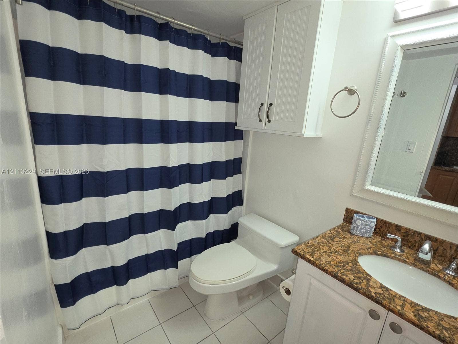 Bathroom featuring white wall-mounted cabinetry, a stone-surfaced vanity with an integrated oval sink, a wall-mounted mirror with decorative molding, and tile flooring