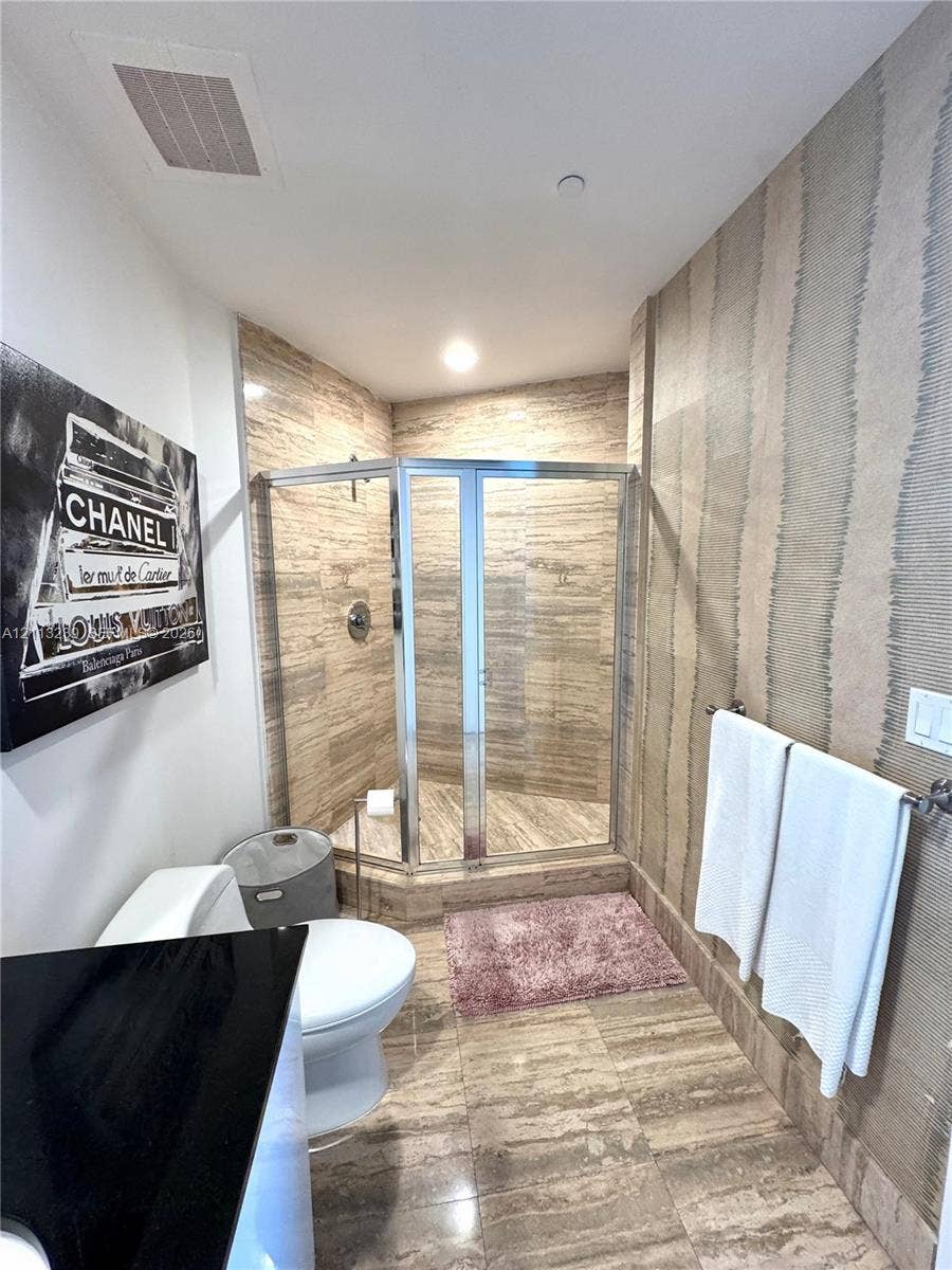16001 Collins Avenue, #2403, Sunny Isles Beach, Sunny Isles Beach, FL 33160 Bathroom featuring a corner glass-enclosed shower, stone tile flooring, stone-tiled shower walls, wall-mounted metallic towel bar, and textured wall treatment