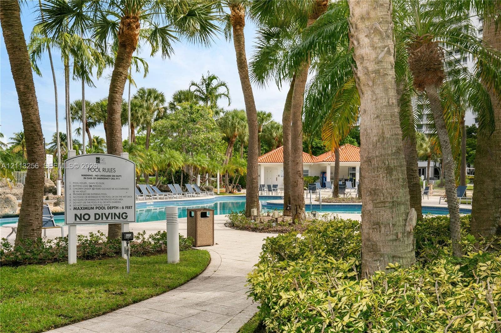 3300 NE 191st Street, #1514, Aventura, FL 33180 Swimming pool area featuring a stone-paved deck, mature palm trees, landscaped shrubbery, and a covered structure with a terracotta tile roof
