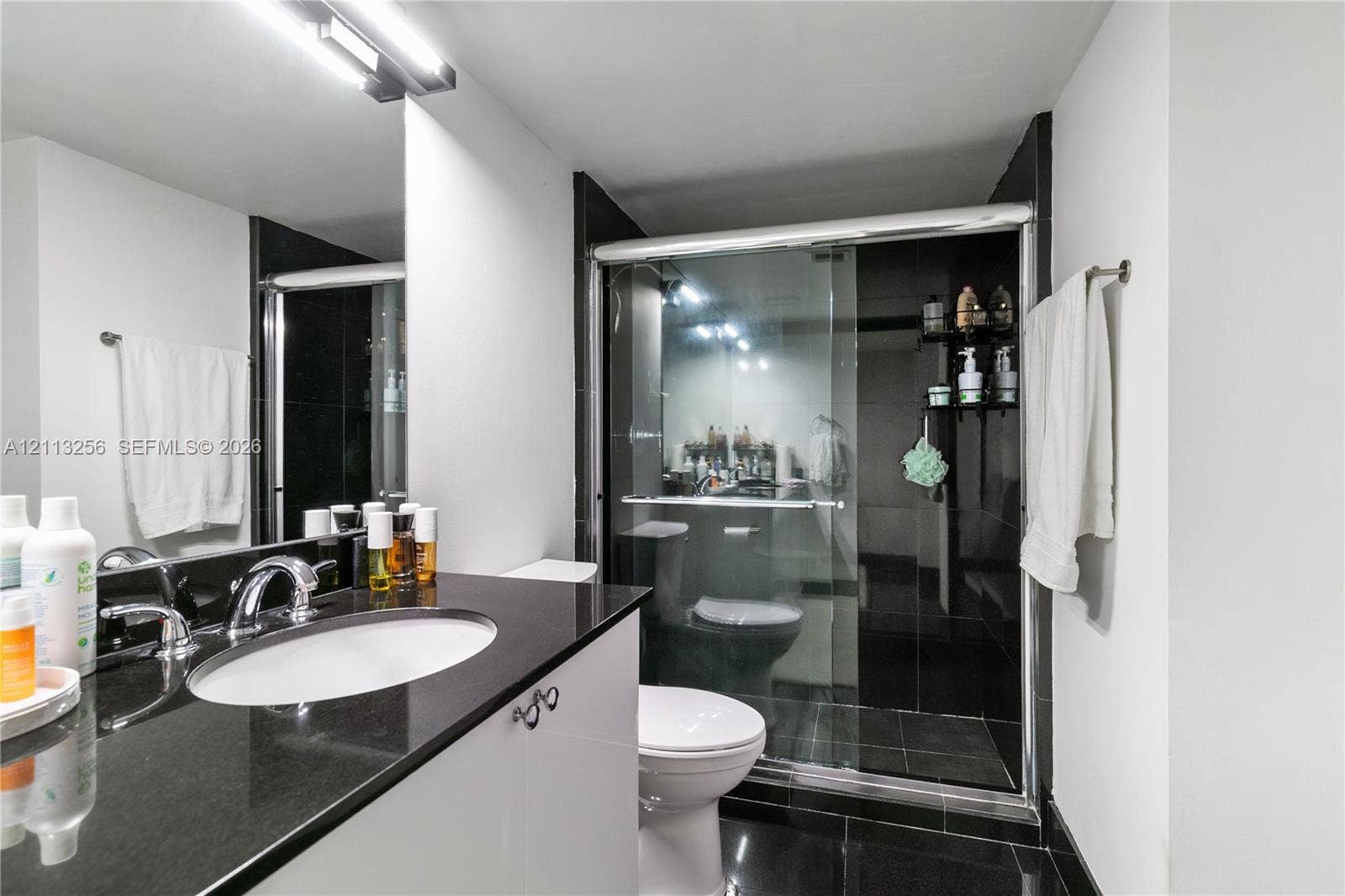 3300 NE 191st Street, #1514, Aventura, FL 33180 Bathroom featuring a dark stone countertop, dual sinks, white cabinetry, a glass-enclosed shower with dark tile surround, and black tiled flooring