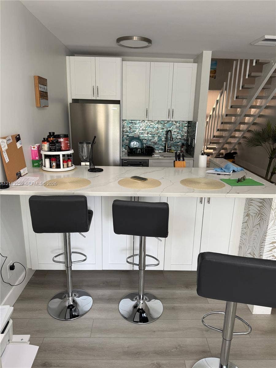 Kitchen featuring white shaker-style cabinetry, a stone-finish countertop peninsula, mosaic tile backsplash, stainless steel refrigerator, and wood-finish flooring